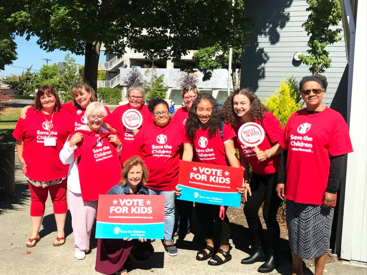 Group of volunteers wearing red Save the Children shirts holding “Vote for Kids” signs at an outdoor community event.
